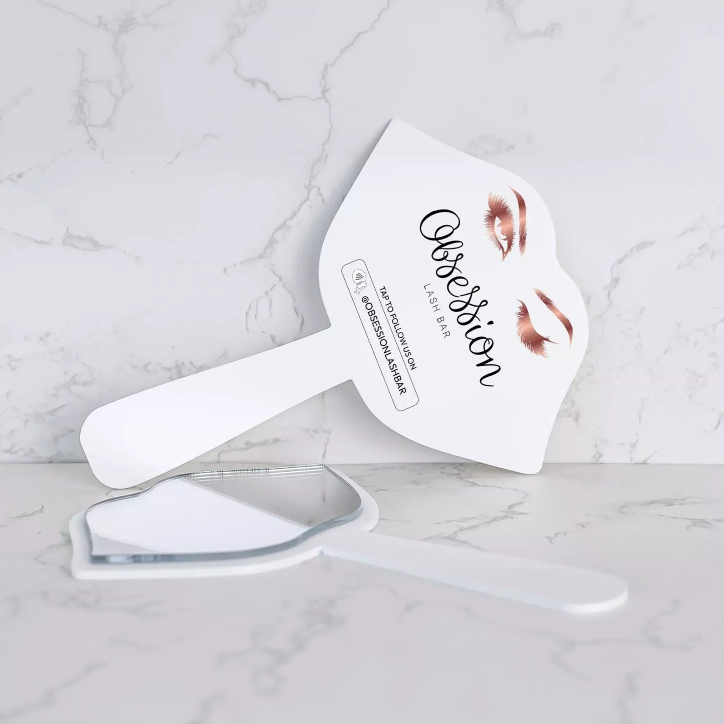 A white makeup brush with a mirror attached to it, placed on a marble surface.